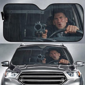 May include: A rectangular car windshield sunshade featuring a printed graphic of a man driving a vehicle while holding a firearm. The sunshade is black with a matte finish and is designed to cover the front windshield of a car to block sunlight.
