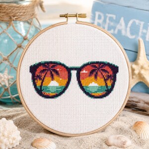 May include: A cross-stitch embroidery piece in a round wooden hoop featuring a pair of sunglasses. The lenses display a colorful sunset scene with silhouettes of palm trees in shades of orange, yellow, purple, and teal against a white fabric.
