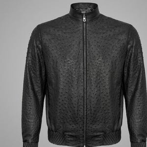 Può includere: Una giacca nera con una superficie testurizzata a pois che ricorda la pelle di struzzo. La giacca presenta un colletto alto, una cerniera centrale a tutta lunghezza con un cursore metallico, polsini e orlo a coste per una vestibilità strutturata.