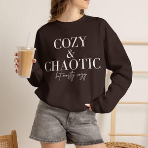 May include: A dark brown long-sleeve crewneck sweatshirt featuring the white text COZY & CHAOTIC but mostly cozy printed on the front. The garment is paired with grey denim shorts. The design uses a mix of bold serif and script fonts.