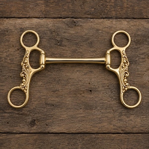 Op de afbeelding: Een goudkleurig metalen paardenbit met een recht mondstuk en twee zijstukken. Elk zijstuk is versierd met een gedetailleerd, verhoogd krulpatroon en is voorzien van twee ronde ringen aan de boven- en onderkant voor bevestiging.