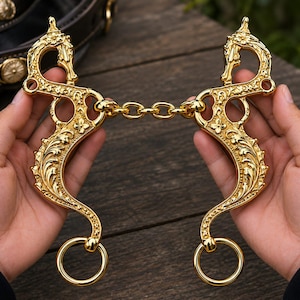 Könnte beinhalten: Ein goldfarbenes Pferdegebiss aus Metall mit zwei verzierten Seitenteilen, die mit detaillierten Blumen- und Rankengravuren versehen sind. Die Seitenteile sind durch eine mittlere Metallkette verbunden und enden jeweils in einem Ring.