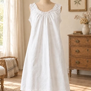 May include: A sleeveless white nightdress featuring a round neckline with delicate ruffled lace trim. The garment has a loose, flowing silhouette that falls to the knee, with subtle gathered detailing at the collar and a small embroidered accent.