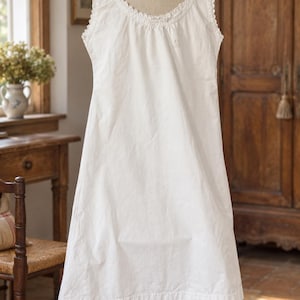 Puede incluir: Un camisón blanco sin mangas que presenta un cuello redondo con un delicado borde de encaje y un pequeño detalle bordado en la parte superior del pecho. La prenda tiene una silueta holgada y fluida que llega hasta las rodillas.