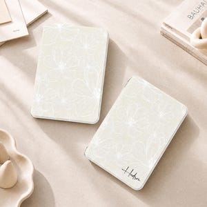 Könnte beinhalten: Zwei rechteckige, hellbeige Etuis mit einem zarten, weißen Linienmuster aus Blumen. Das rechte Etui trägt den Namen Hudson in einer schwarzen, kursiven Schriftart. Beide Gegenstände haben abgerundete Ecken und eine glatte, matte Oberfläche.