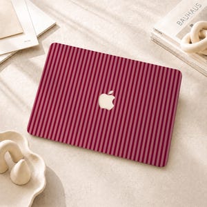 Peut inclure: Un autocollant pour ordinateur portable avec un motif &agrave; rayures verticales alternant des nuances de bordeaux profond et de rose clair. Le design pr&eacute;sente une d&eacute;coupe centrale r&eacute;v&eacute;lant le logo Apple blanc sur le capot de l'ordinateur.