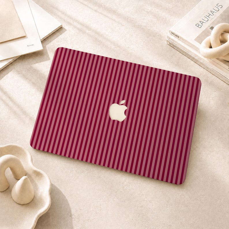 Peut inclure: Un autocollant pour ordinateur portable avec un motif &agrave; rayures verticales alternant des nuances de bordeaux profond et de rose clair. Le design pr&eacute;sente une d&eacute;coupe centrale r&eacute;v&eacute;lant le logo Apple blanc sur le capot de l'ordinateur.