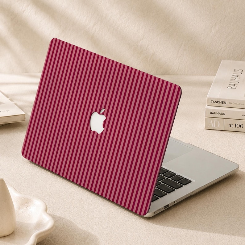 Peut inclure: Un ordinateur portable dot&eacute; d'une coque de protection &agrave; rayures verticales dans des tons rouge fonc&eacute; et rose clair. Le logo pomme blanc embl&eacute;matique est visible au centre du couvercle, tandis que le clavier et le pav&eacute; tactile argent&eacute;s sont visibles.