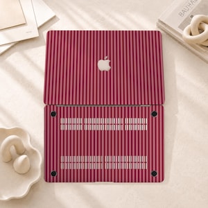 Peut inclure: Une coque rigide pour ordinateur portable avec un motif &agrave; rayures verticales dans des tons altern&eacute;s de rouge fonc&eacute; et de rose clair. La partie sup&eacute;rieure pr&eacute;sente une d&eacute;coupe centrale pour le logo Apple, tandis que la base comporte des fentes d'a&eacute;ration.