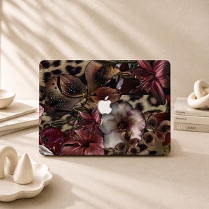 Peut inclure: Une coque rigide rectangulaire pour ordinateur portable ornée d'un imprimé floral composé de lys rouge foncé, d'hibiscus et d'une cerise sur un fond léopard beige et noir, avec une découpe centrale pour le logo Apple argenté.