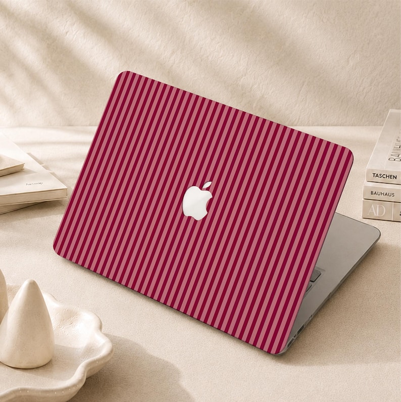 Peut inclure: Un ordinateur portable dot&eacute; d'une coque de protection &agrave; rayures verticales alternant des tons rouge fonc&eacute; et rose clair. Le logo Apple blanc est visible au centre du couvercle, qui est ferm&eacute; et pos&eacute; sur une surface plane.