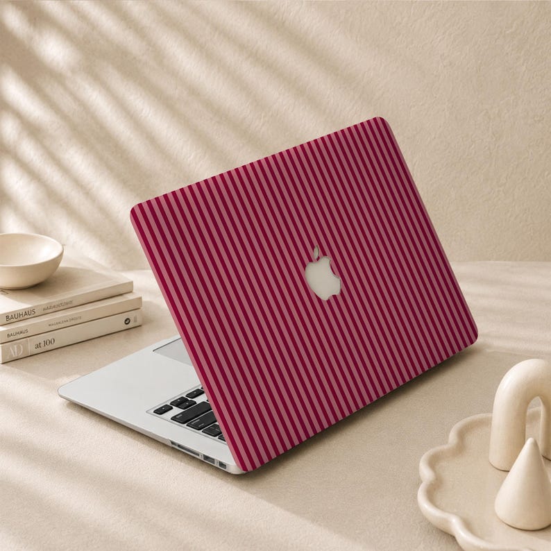 Peut inclure: Un ordinateur portable avec une coque de protection arborant un motif de rayures verticales rouge fonc&eacute; et rose clair. Le logo Apple est visible au centre du couvercle. L'ordinateur est ouvert, r&eacute;v&eacute;lant un clavier argent&eacute; et un cadre m&eacute;tallique.