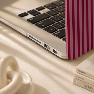 Peut inclure: Un ordinateur portable argent&eacute; dot&eacute; d'une coque de protection &agrave; rayures verticales alternant des tons rose fonc&eacute; et rose clair. L'ordinateur est ouvert, r&eacute;v&eacute;lant un clavier noir avec des lettres blanches, ainsi que des ports USB et lightning.