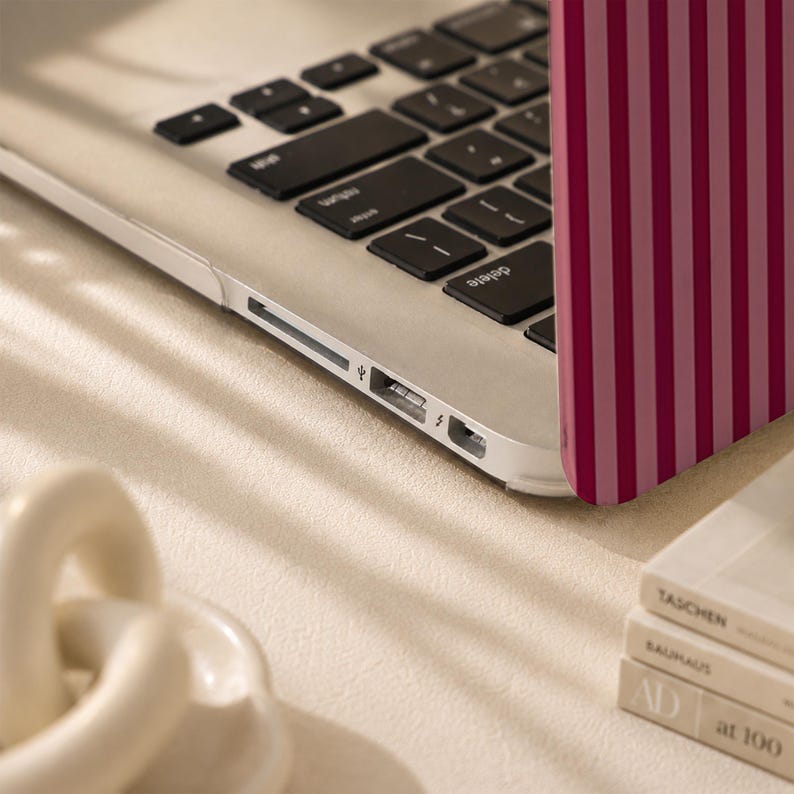 Peut inclure: Un ordinateur portable argent&eacute; dot&eacute; d'une coque de protection &agrave; rayures verticales alternant des tons rose fonc&eacute; et rose clair. L'ordinateur est ouvert, r&eacute;v&eacute;lant un clavier noir avec des lettres blanches, ainsi que des ports USB et lightning.