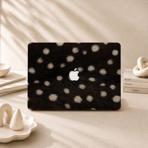 May include: A laptop skin featuring a dark brown and black fur-like texture with scattered white spots. The design covers the exterior lid of a closed laptop, which displays a centred, white, glowing Apple logo. The pattern mimics a natural animal hide.