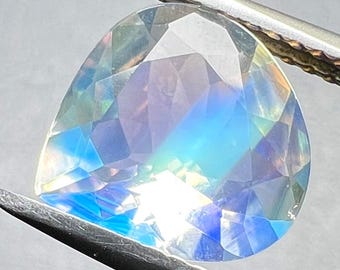 Puede incluir: Una piedra preciosa facetada en forma de pera que muestra destellos iridiscentes de luz azul, morada y blanca. La piedra translúcida está sujeta por unas pinzas de metal plateado, mostrando un corte brillante con múltiples facetas.