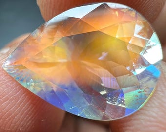 Puede incluir: Una piedra preciosa facetada en forma de pera que muestra un vibrante juego de colores con tonos iridiscentes de naranja, amarillo y azul. La piedra presenta un cuerpo transparente con numerosas facetas geométricas que reflejan la luz.