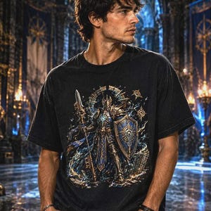 May include: A black short-sleeved t-shirt featuring a detailed graphic print of a knight in ornate armor holding a tall spear and a shield. The design incorporates blue and gold accents, intricate patterns, and celestial elements surrounding the central figure, set against a dark fabric background.