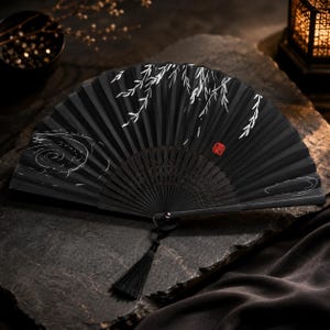 Peut inclure: Un éventail pliant noir orné de motifs de branches de saule peints en blanc et d'un dessin de lignes tourbillonnantes. L'éventail comprend un petit sceau carré rouge sur le côté droit et un pompon en tissu noir fixé à la base du manche.