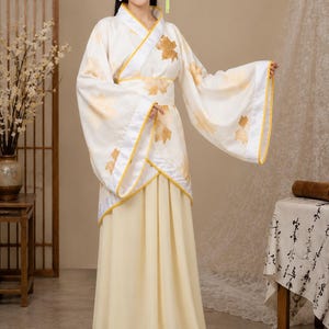 Peut inclure: Un haut blanc style cache-cœur à manches longues et larges avec un liseré jaune, orné d'un motif de feuilles d'érable dorées. Il est associé à une jupe longue de couleur jaune pâle uni et à une large ceinture assortie avec des détails de feuilles.