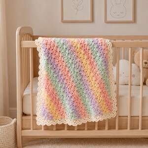 Puede incluir: Una manta de ganchillo rectangular para bebé con rayas diagonales en tonos pastel de rosa, amarillo, verde y morado. La manta presenta un borde festoneado de color crema y un patrón de puntos texturizado en toda la pieza.