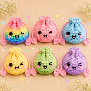 May include: Six round, crocheted drawstring pouches arranged in two rows. Each pouch features a smiling face with black eyes, a small mouth, and pink cheeks. The pouches are colored rainbow, pink, blue, yellow, green, and purple with leaf-shaped ends.
