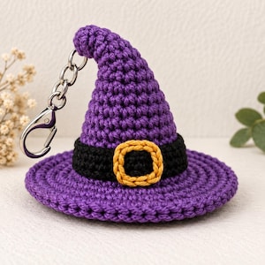 Puede incluir: Un llavero de sombrero de bruja tejido a ganchillo en color morado con una banda negra en la base y una hebilla cuadrada amarilla. El sombrero tiene una punta ligeramente curvada y un ala ancha. Un mosquetón de metal plateado está sujeto al lado.