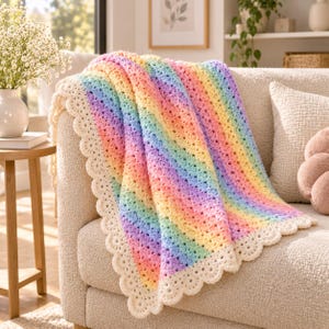 Puede incluir: Una manta de ganchillo con rayas diagonales en un suave degradado arcoíris de tonos pastel rosa, amarillo, azul y morado. La manta está rematada con un borde ancho festoneado de color crema que enmarca la parte central colorida.