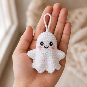 May include: A small white crochet ghost ornament featuring a rounded head, two outstretched arms, and a scalloped bottom edge. The ghost has black embroidered eyes with white highlights, a black stitched smile, and two small pink circular cheeks.