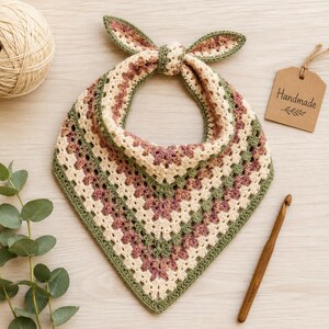 Pode incluir: Uma bandana triangular de crochê com padrão granny square em listras alternadas de creme, verde sálvia e rosa antigo. O acessório inclui dois laços superiores para amarrar e uma etiqueta marrom com a inscrição Handmade.