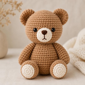 May include: A small, handcrafted crochet teddy bear made with light brown yarn. The bear features a round head, two small ears, a cream-coloured muzzle with a dark brown embroidered nose and mouth, and two black plastic eyes. The feet have cream circles.