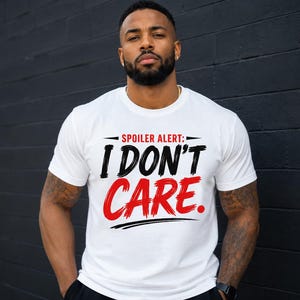 Könnte beinhalten: Ein weißes kurzärmeliges T-Shirt mit dem Text SPOILER ALERT: I DON'T CARE. Die Worte SPOILER ALERT sind in kleinen schwarzen Buchstaben, I DON'T in einer großen schwarzen Pinselstrich-Schrift und CARE in einer großen roten Pinselstrich-Schrift.