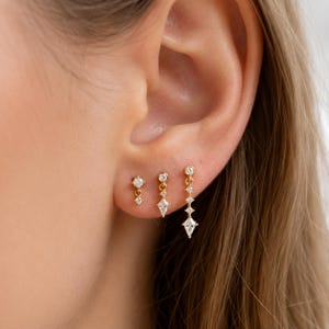 May include: Three gold-toned drop earrings arranged in a row on an earlobe. Each earring features small, clear, sparkling stones set in metal. The first is a short drop, the second is a medium drop with a kite-shaped stone, and the third is a longer drop.