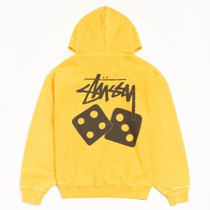Puede incluir: Una sudadera con capucha de color amarillo brillante que presenta un gran gráfico negro en la parte posterior. El diseño incluye el logotipo de Stüssy en una fuente manuscrita estilizada sobre dos dados negros, uno con cuatro puntos y otro con tres.