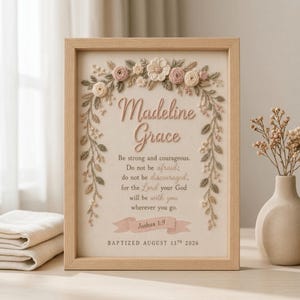 May include: A framed wall art piece featuring embroidered floral accents in shades of pink, cream, and green surrounding the name Madeline Grace and the Bible verse Joshua 1:9, with the text Baptized August 13th 2026 printed at the bottom.