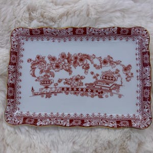 May include: A rectangular porcelain tray with a white base and a red floral pattern. The centre features a detailed illustration of flowers and vases. The border is decorated with a repeating floral motif and finished with a thin gold-coloured rim.