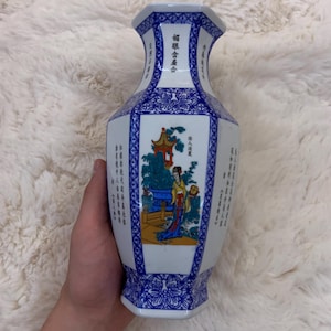 Può includere: Un vaso esagonale in porcellana con base bianca e intricati motivi floreali e geometrici blu. Il pannello centrale mostra un'illustrazione colorata di una figura in abiti tradizionali vicino a un padiglione, accompagnata da testo cinese nero.