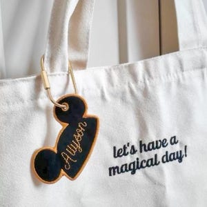K&ouml;nnte beinhalten: Eine wei&szlig;e Canvas-Tragetasche mit dem gestickten schwarzen Text "let's have a magical day!" und einem schwarzen, mausohr-f&ouml;rmigen Kofferanh&auml;nger mit goldenem Rand und dem Namen "Alyson" in goldener Schreibschrift, befestigt mit einem goldenen Karabiner.