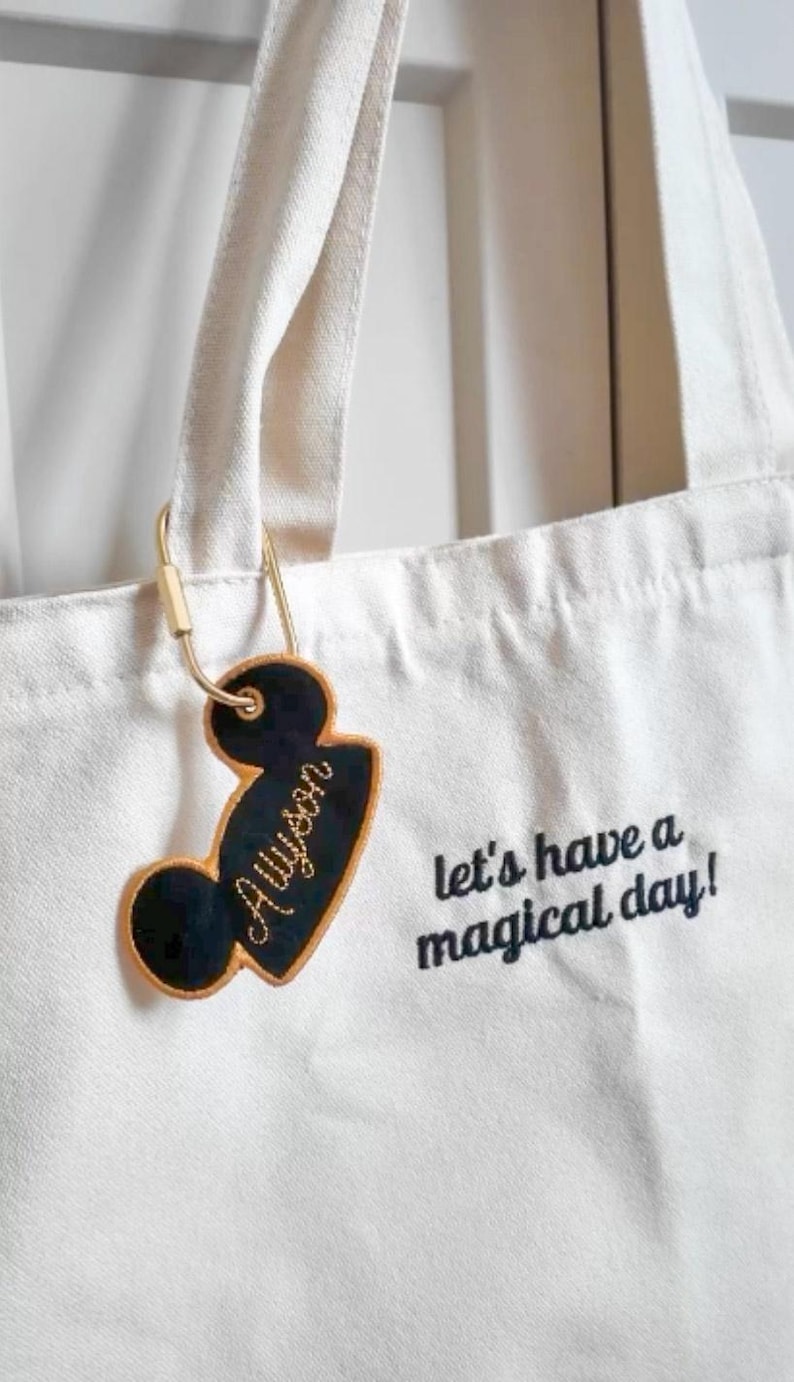 K&ouml;nnte beinhalten: Eine wei&szlig;e Canvas-Tragetasche mit dem gestickten schwarzen Text "let's have a magical day!" und einem schwarzen, mausohr-f&ouml;rmigen Kofferanh&auml;nger mit goldenem Rand und dem Namen "Alyson" in goldener Schreibschrift, befestigt mit einem goldenen Karabiner.