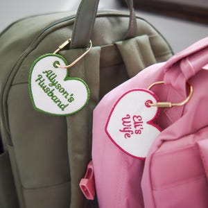 May include: Two heart-shaped bag tags attached to backpacks with gold-tone metal clips. One white tag with green trim and green embroidery reads Allyson's Husband. The other white tag with pink trim and pink embroidery reads Eli's Wife.