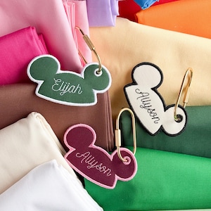 May include: Three mouse-ear shaped fabric tags with gold-toned metal carabiners. One tag is dark green with white embroidery reading Elijah, one is cream with black embroidery reading Allyson, and one is maroon with pink embroidery reading Allyson.