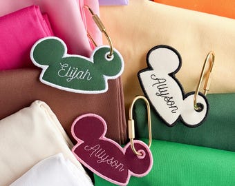 Könnte beinhalten: Drei mausohr-förmige Stoffanhänger mit goldfarbenen Metallkarabinern. Ein Anhänger ist dunkelgrün mit weißer Stickerei Elijah, einer cremefarben mit schwarzer Stickerei Allyson und einer kastanienbraun mit rosa Stickerei Allyson.