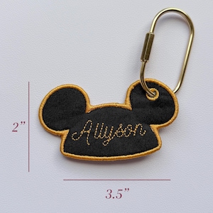 K&ouml;nnte beinhalten: Ein schwarzer, bestickter Schl&uuml;sselanh&auml;nger in Form von Mausohren mit goldener Stickerei und dem Namen Allyson in Schreibschrift. Er verf&uuml;gt &uuml;ber einen goldfarbenen Metallkarabiner. Der Anh&auml;nger ist 5,08 cm hoch und 8,89 cm breit, wie abgebildet.