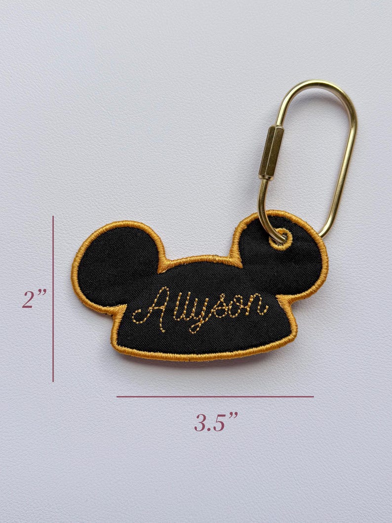 K&ouml;nnte beinhalten: Ein schwarzer, bestickter Schl&uuml;sselanh&auml;nger in Form von Mausohren mit goldener Stickerei und dem Namen Allyson in Schreibschrift. Er verf&uuml;gt &uuml;ber einen goldfarbenen Metallkarabiner. Der Anh&auml;nger ist 5,08 cm hoch und 8,89 cm breit, wie abgebildet.