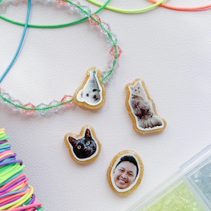 May include: A collection of custom resin charms featuring photographic prints of two cats, one dog wearing a party hat, and a smiling person. The charms have a gold glitter border and are displayed alongside a beaded necklace and colorful elastic cords.