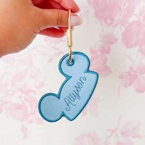 May include: A light blue embroidered keychain shaped like a mouse head with a dark blue stitched border. The name Allyson is embroidered in dark blue cursive script across the center. A gold-toned metal ring is attached to the top eyelet.