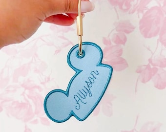 Könnte beinhalten: Ein hellblauer, bestickter Schlüsselanhänger in Form eines Mauskopfes mit dunkelblauem Stickrand. Der Name Allyson ist in dunkelblauer Schreibschrift in die Mitte gestickt. Ein goldfarbener Metallring ist an der oberen Öse befestigt.