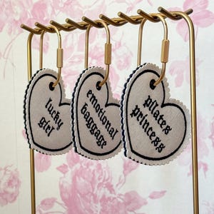 May include: Three heart-shaped fabric tags with dark blue scalloped edges and black embroidered text hanging from a gold metal stand. The tags feature the phrases "lucky girl," "emotional baggage," and "pilates princess" in a gothic font.