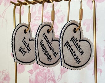 Könnte beinhalten: Drei herzförmige Stoffanhänger mit dunkelblauem Wellenrand und schwarzer Stickerei hängen an einem goldfarbenen Metallständer. Die Anhänger tragen die Aufschriften "lucky girl", "emotional baggage" und "pilates princess" in gotischer Schrift.