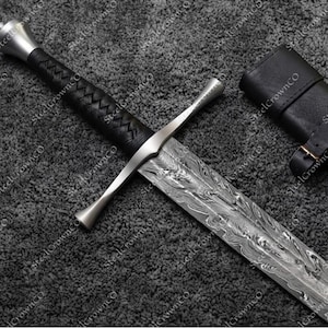 Op de afbeelding: Een zwaard van Damascus-staal met een zilverkleurige pareerstang en pommel, voorzien van een zwart lederen handvat met een kruispatroon. Het lemmet vertoont een opvallend golvend patroon. Een zwarte lederen schede met riemen ligt naast het zwaard.